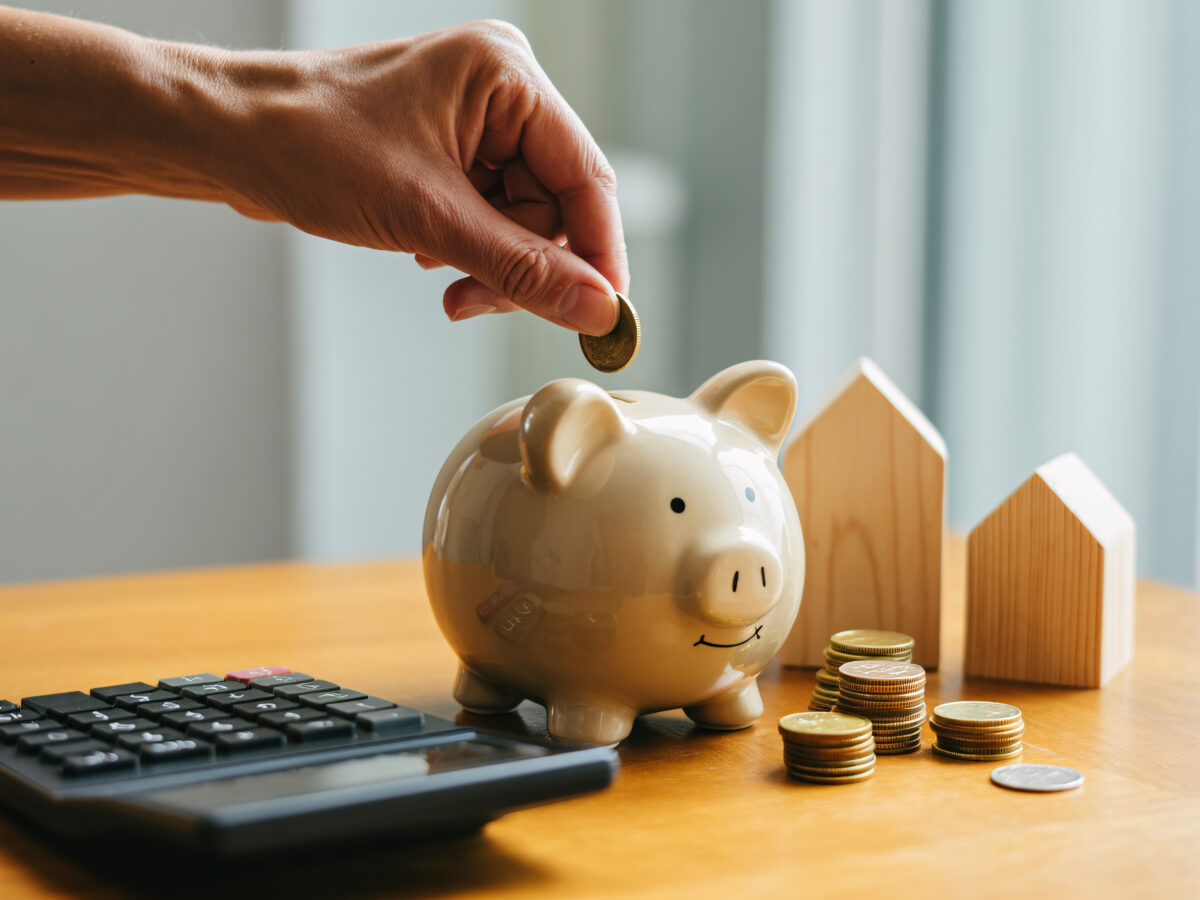 A person's hand is shown dropping a gold coin into a ceramic piggy bank. Next to the piggy bank are stacks of gold coins and three small wooden house models. A calculator is positioned on the left side of the wooden table. The background features a blurred window with vertical blinds.