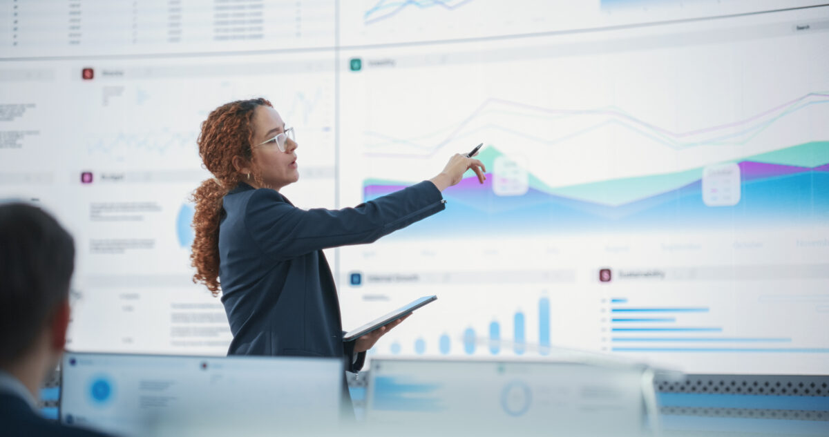 Female Senior Data Scientist Reviewing Reports Of Risk Management Department On Big Digital Screen In Monitoring Room. Diverse Consulting Company Employees Working Behind Desktop Computers.