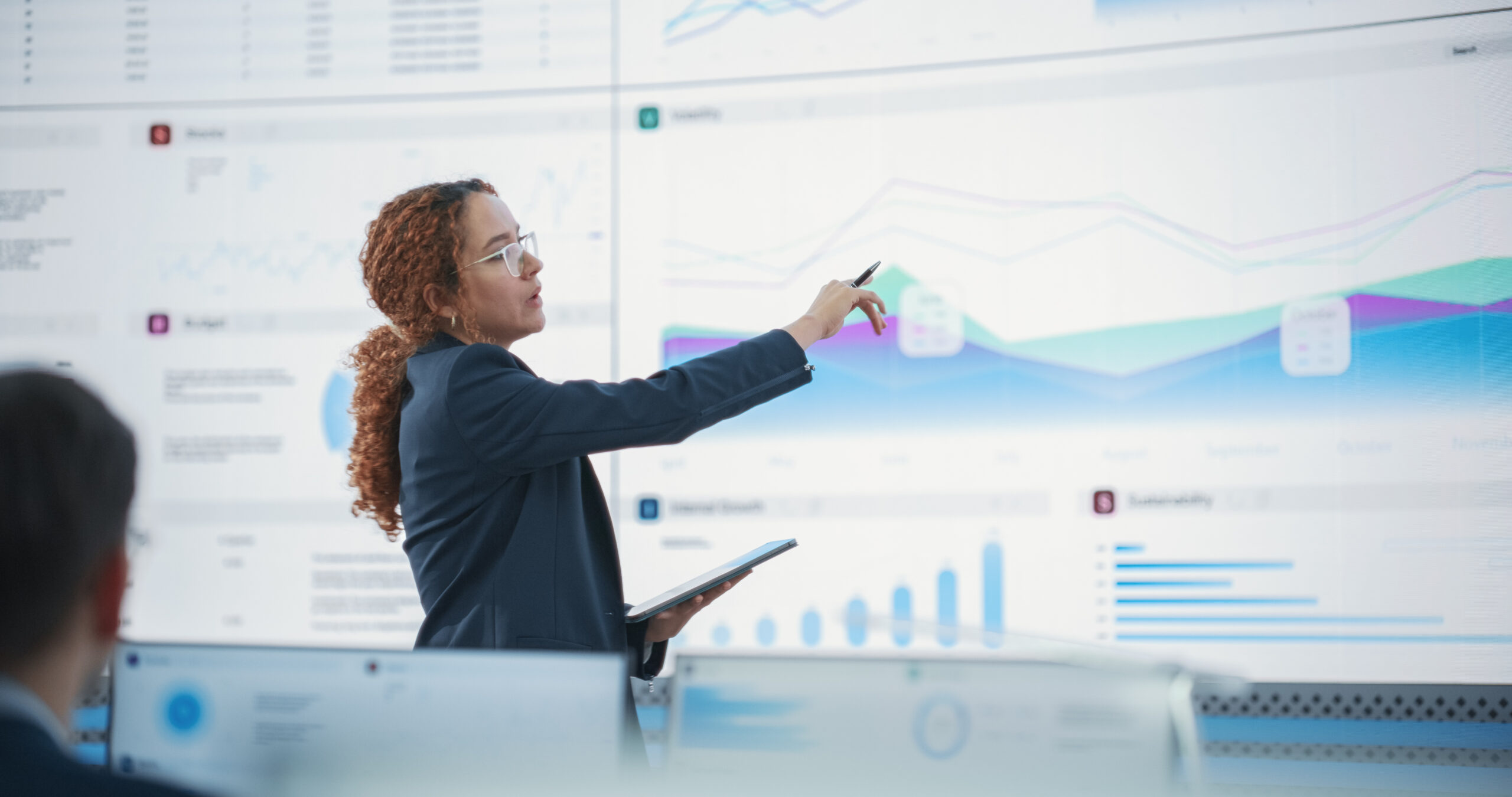 Female Senior Data Scientist Reviewing Reports Of Risk Management Department On Big Digital Screen In Monitoring Room. Diverse Consulting Company Employees Working Behind Desktop Computers.